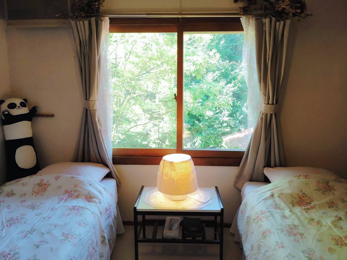 A room with two beds, a window with curtains, a table with a lamp, and a panda decoration.