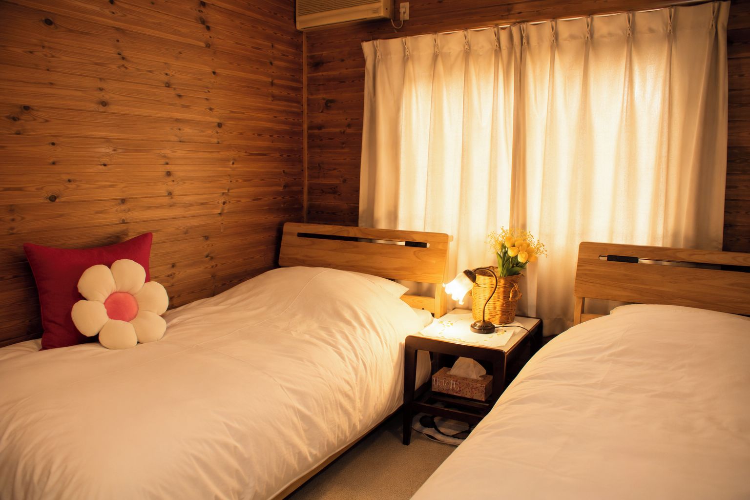 A room with two beds, wooden walls, a nightstand with a lamp, and a window with curtains.