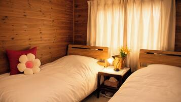 A room with two beds, wooden walls, a nightstand with a lamp, and a window with curtains.