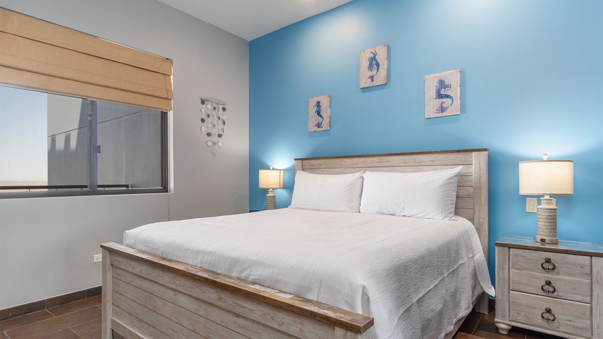 Een slaapkamer met een houten bed, twee nachtkastjes met lampen, een raam met zonwering en drie aan de muur hangende kunstwerken.