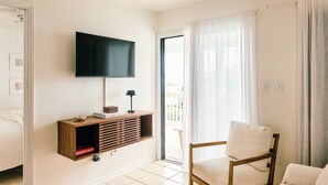 Ein Wohnzimmer mit einem an der Wand befestigten Flachbildfernseher, einer Holzunterhaltungsecke darunter, einem weißen Sessel und einer Glastür, die zu einem Außenbereich führt.