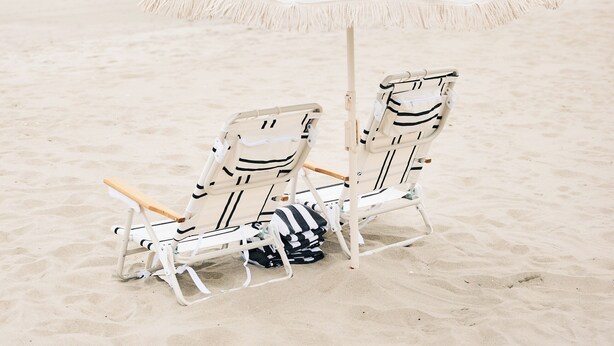 Zwei Liegestühle unter einem weißen Sonnenschirm an einem sandigen Strand, im Hintergrund das Meer.