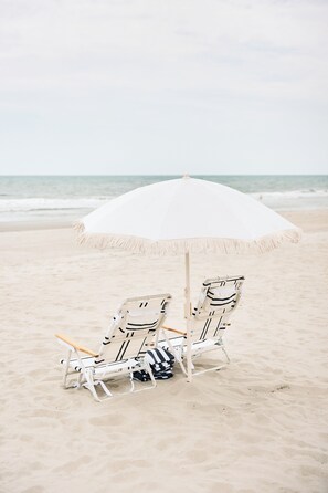 Deux chaises de plage sous un parasol blanc, sur une plage de sable, avec l’océan en arrière-plan.