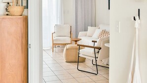 Ein geräumiges Wohnzimmer mit einem weißen Sofa, einem Holztisch und einem Sessel. Ein Deckenventilator ist zu sehen, und es gibt ein Fenster mit Vorhängen.
