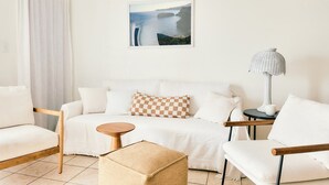 Ein modernes, minimalistisches Zimmer mit einem weißen Sofa, einem Holztisch und einem gewebten Hocker. Ein gerahmtes Bild einer Küstenlandschaft hängt an der Wand.
