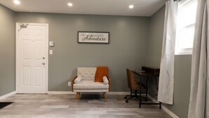 A room with a grey wall, a white door, a beige armchair with an orange throw, a wooden desk, and a white curtain.