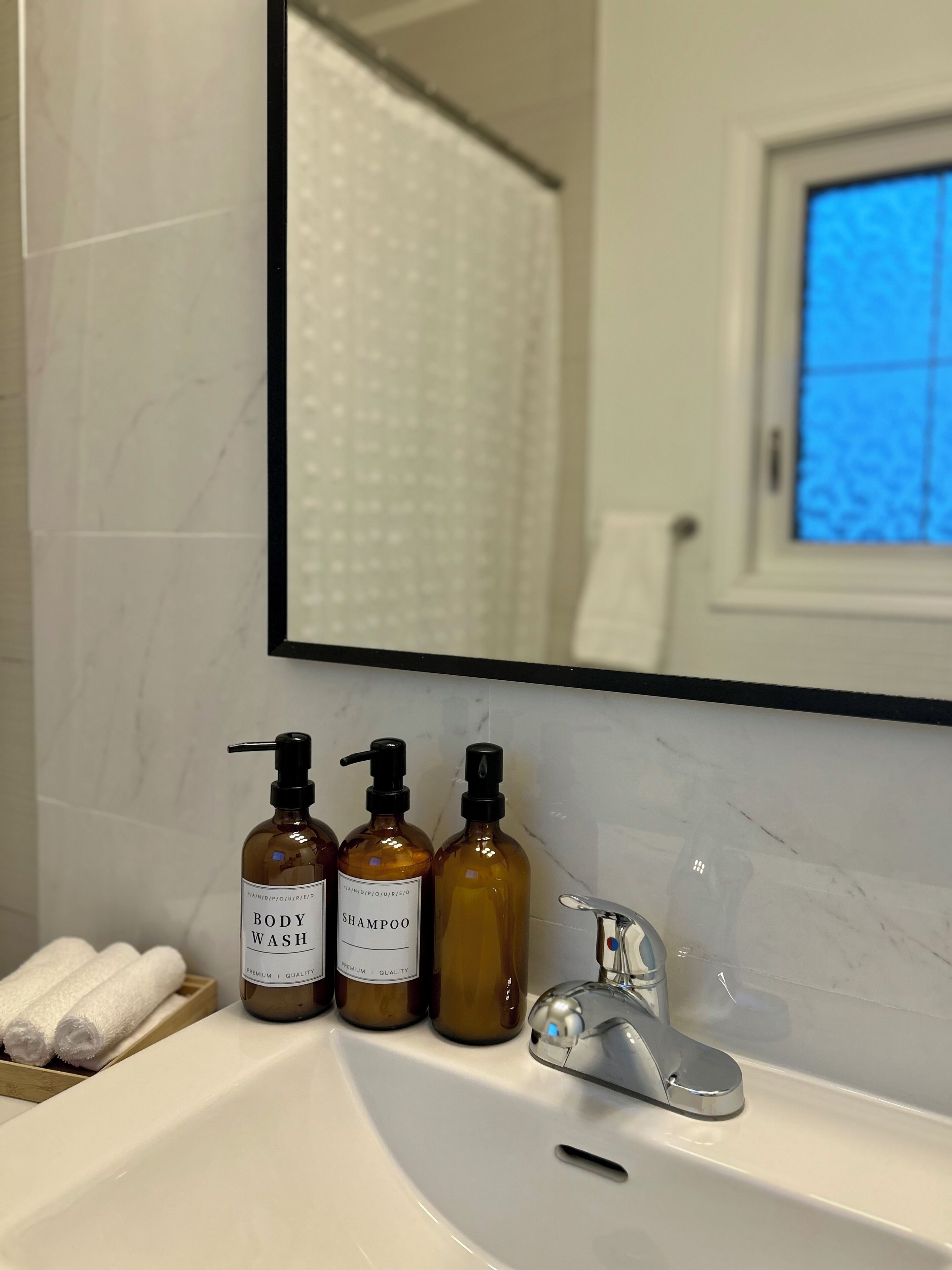 Een badkamer met een wit wastafel, twee flessen douchegel en een spiegel die een raam met blauwe lucht weerspiegelt.