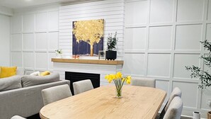 A modern living room with a wooden dining table, grey upholstered chairs, a sofa with yellow cushions, a white wall with a grid pattern, and a painting above the fireplace. - Lovelyhome•Near BU•With TreeHouse•Ideal for group stays (Brandon)