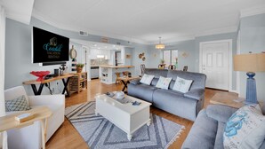 Ein geräumiges Wohnzimmer mit grauem Sofa, weißem Couchtisch und einem Fernseher, der 'Coastal Vacation Rentals' anzeigt. Im Hintergrund ist eine Küche mit weißen Schränken und einer Holz Arbeitsplatte zu sehen.