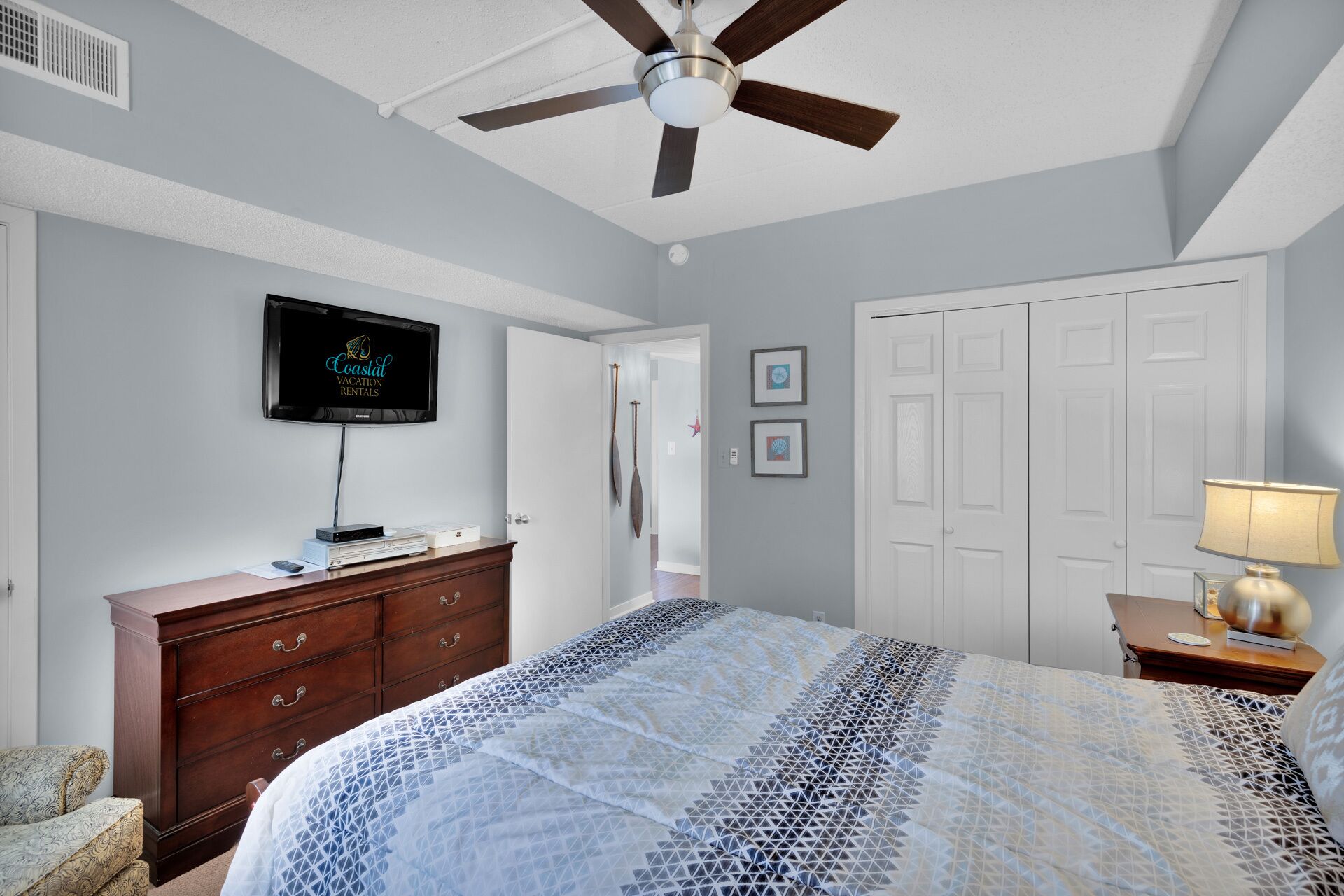 A bedroom with a bed, dresser, TV, ceiling fan, and a door.