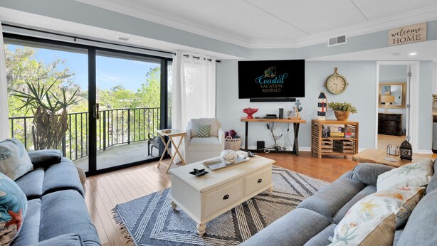 Ein großzügiges Wohnzimmer mit einer großen Schiebeglastür, die auf einen Balkon führt, einem an der Wand befestigten Flachbildfernseher und einer gemütlichen Sitzecke mit Sofa und Couchtisch.