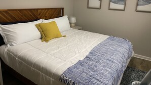 Ein ordentlich bezogenes Bett mit weißer Daunendecke, einem senfgelben Kissen und einer blau-weißen Decke mit Muster. An der Wand hängen drei gerahmte Walbilder.