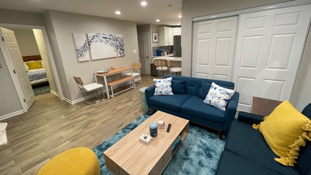 Ein modernes Wohnzimmer mit einem blauen Sofa, einem Holztisch und einem gelben Hocker. Ein Essbereich mit Tisch und Stühlen sowie eine Küche im Hintergrund sind ebenfalls zu sehen.