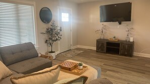 A living room with a grey sofa, a wooden coffee table with a chessboard, a potted plant, and a television mounted on the wall.