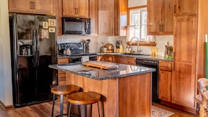 A kitchen with wooden cabinets, a stainless steel refrigerator, a microwave, a stove, and a sink. There are two stools at the island. - The Bartlett Nook/ HOT TUB/ 3-BEDROOM/ 4 BEDS/ CLOSE TO LU (Lynchburg)