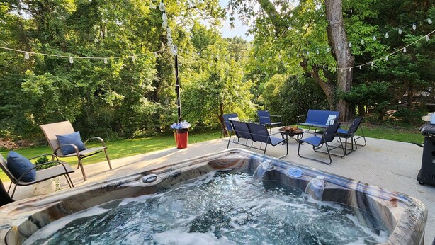 A hot tub surrounded by outdoor furniture, a fire pit, and string lights in a lush garden setting. - The Bartlett Nook/ HOT TUB/ 3-BEDROOM/ 4 BEDS/ CLOSE TO LU (Lynchburg)
