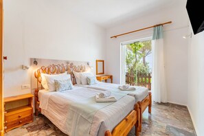 Quarto com cama grande, mesa de cabeceira, janela com vista para árvores e cabeceira de madeira.