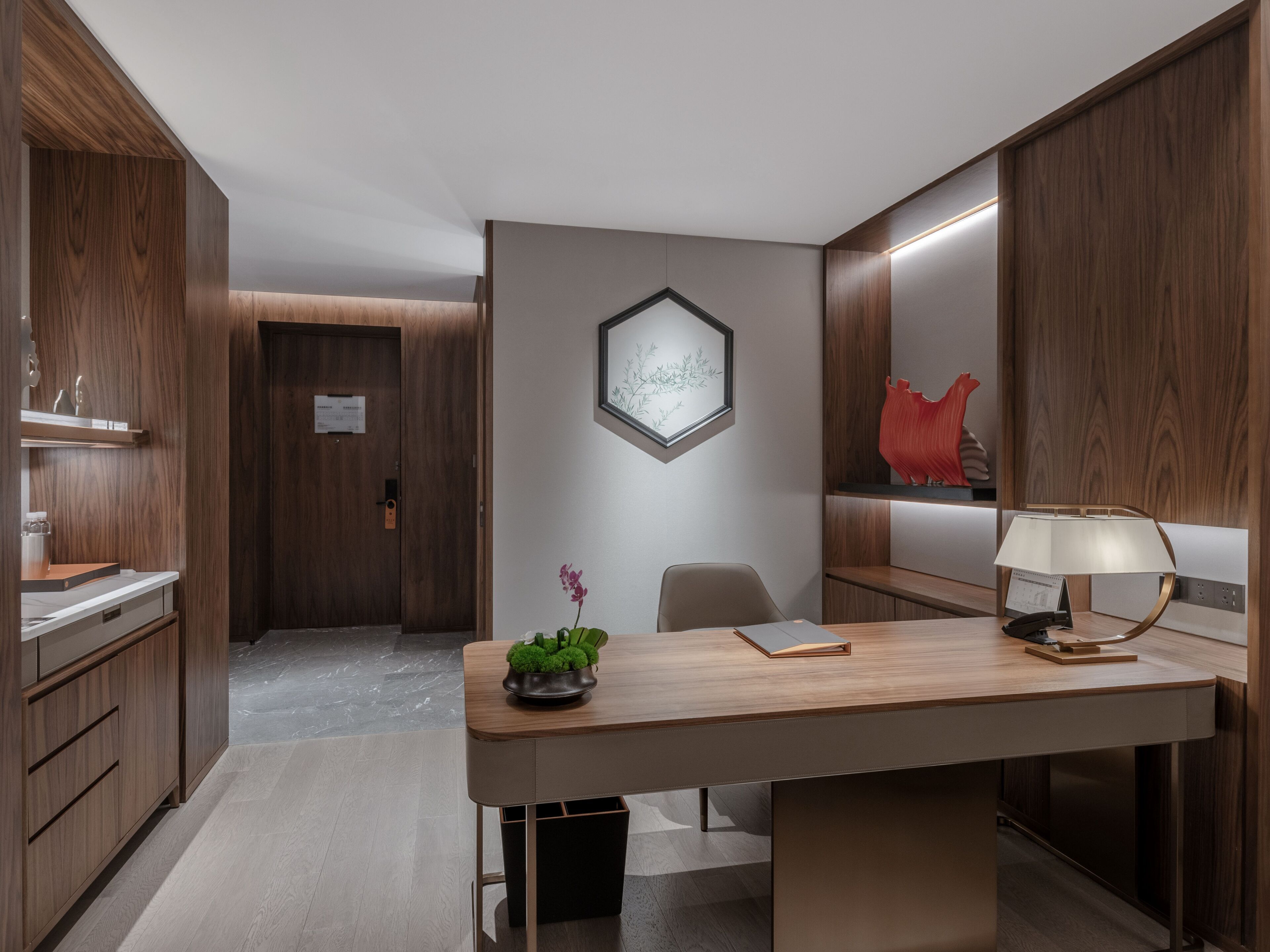 A modern hotel room with a wooden desk, a chair, a small plant, a framed picture, and a red sculpture on the wall.
