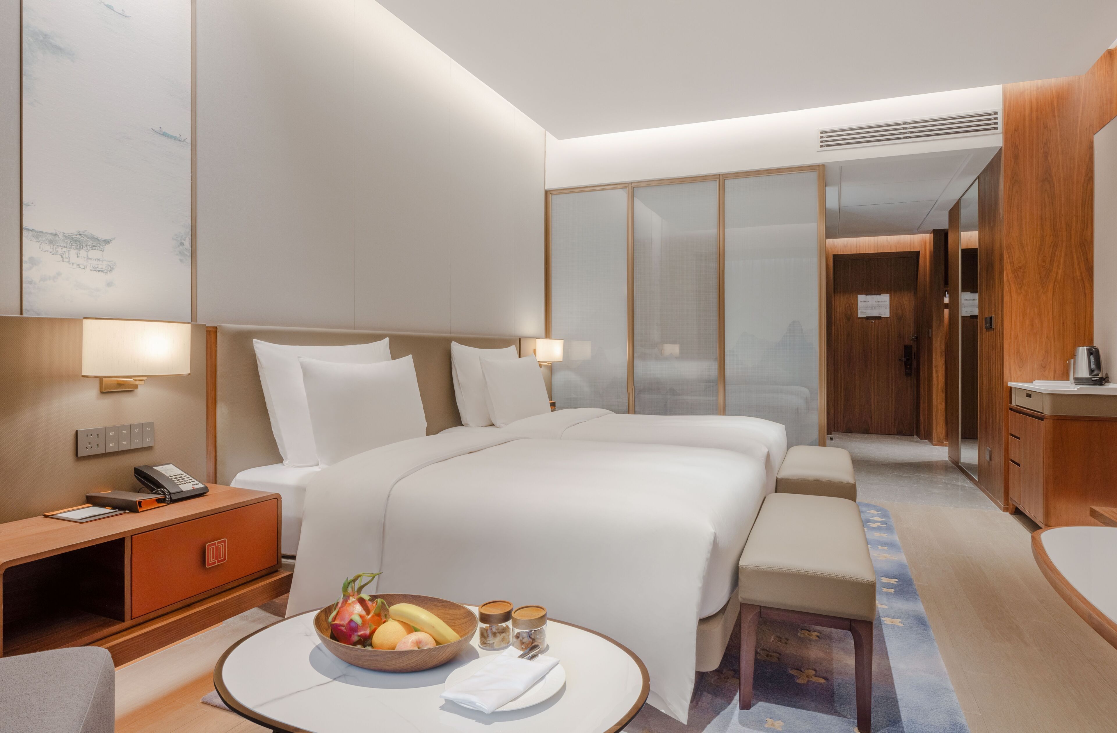 A hotel room with a large bed, bedside tables, a small seating area, a wooden desk, and a fruit bowl.