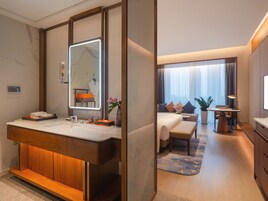 A modern hotel room with a large mirror, a wooden vanity, a bed with a view, a small table, and a desk.