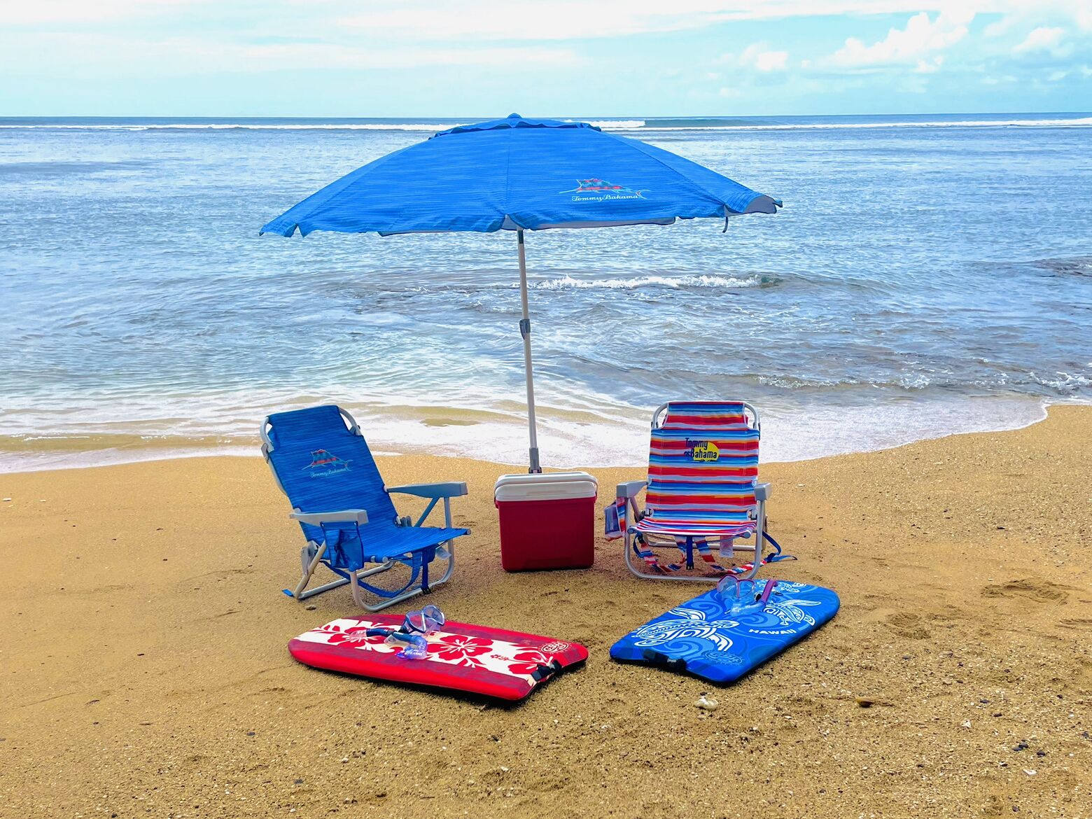 A beach scene with two beach chairs, a blue umbrella, a cooler, and two surfboards on the sand.