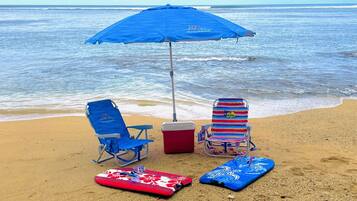 A beach scene with two beach chairs, a blue umbrella, a cooler, and two surfboards on the sand.