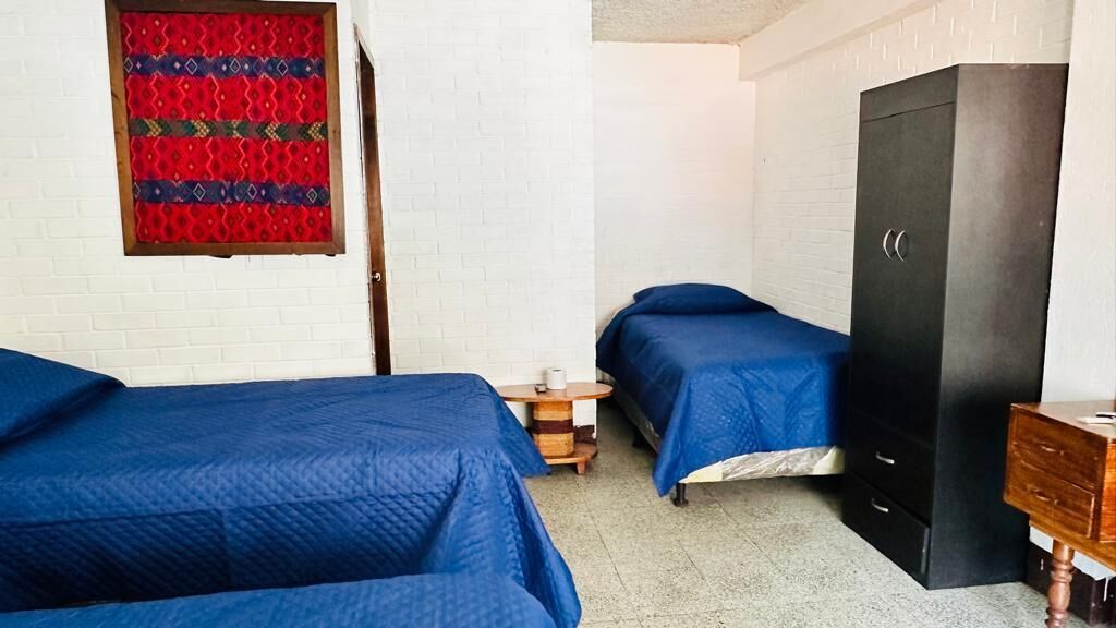 A room with two beds, a wardrobe, a small table, and a wall with a patterned curtain.