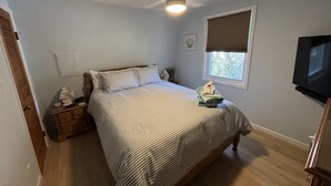 Quarto com cama, mesa de cabeceira, ventilador de teto, janela com persianas e televisão fixada na parede.