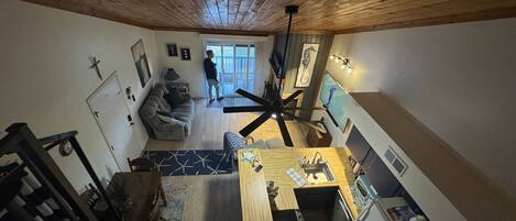A spacious living area with a wooden ceiling, a dining table, a kitchen with a stove and sink, and a large window.