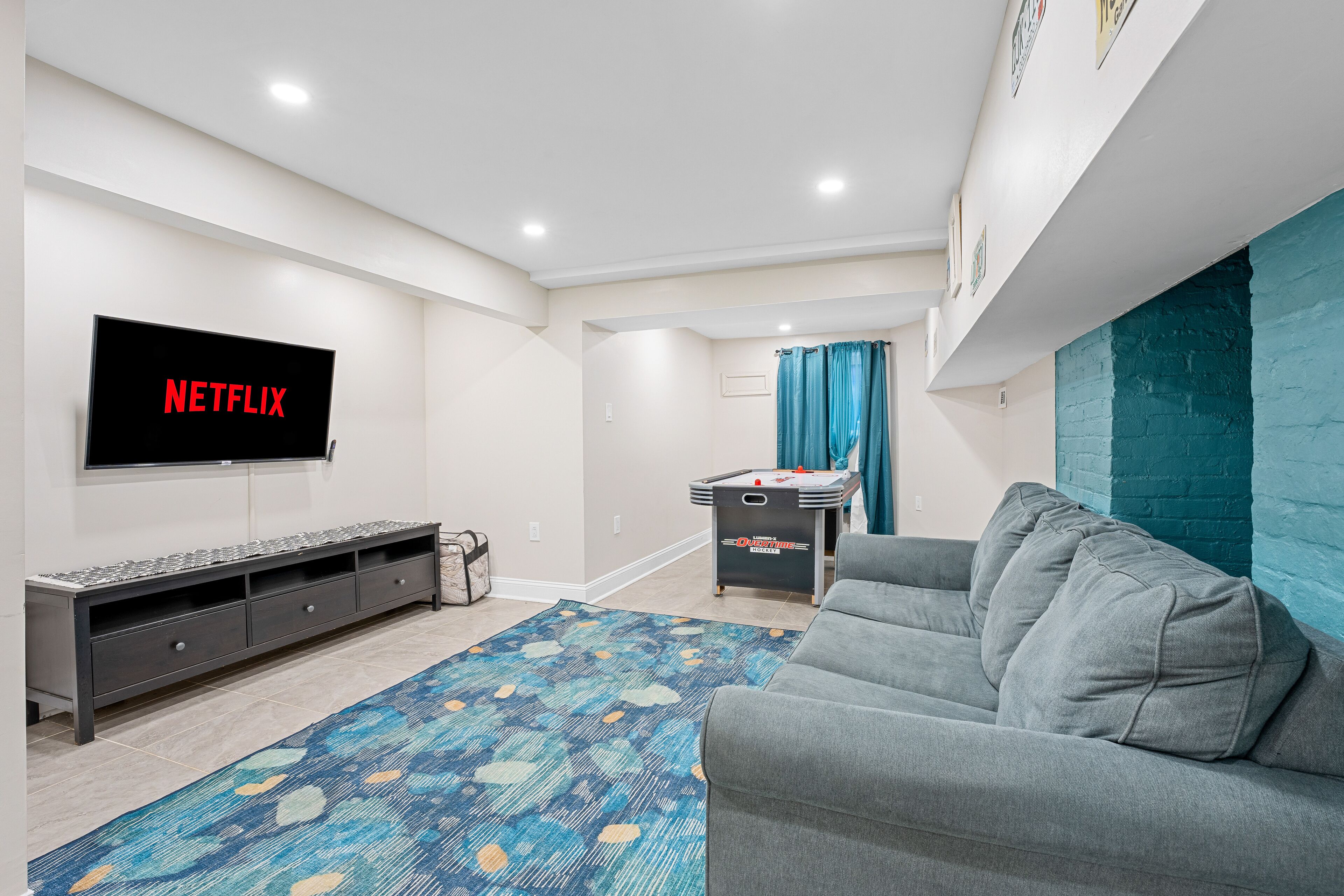 A modern living room with a flat-screen TV, grey sofa, patterned rug, and a pool table.