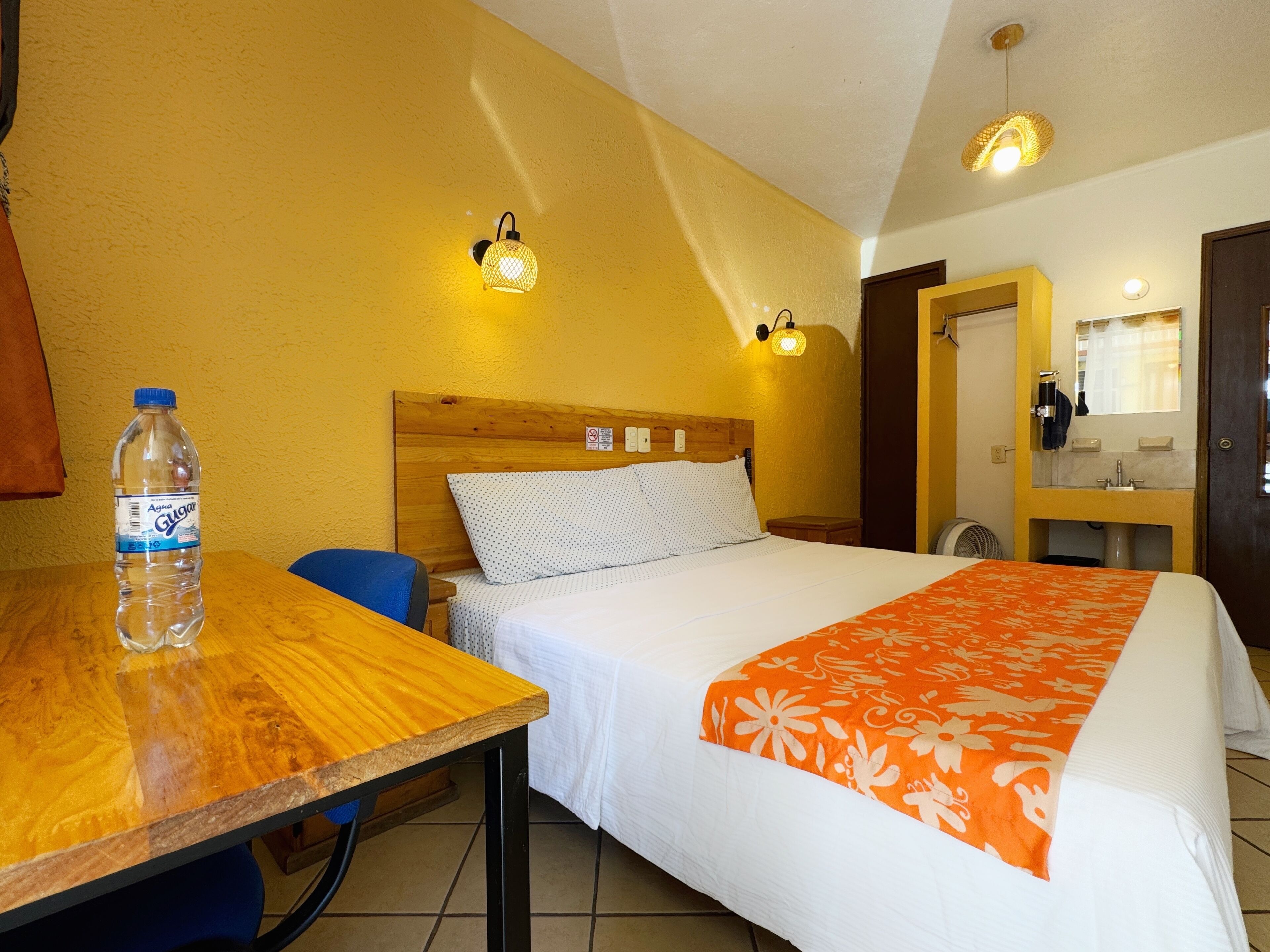 Una habitación de hotel con cabecera de madera, cama con ropa de cama blanca y naranja, una mesa con una botella de agua y dos luces fijadas a la pared.