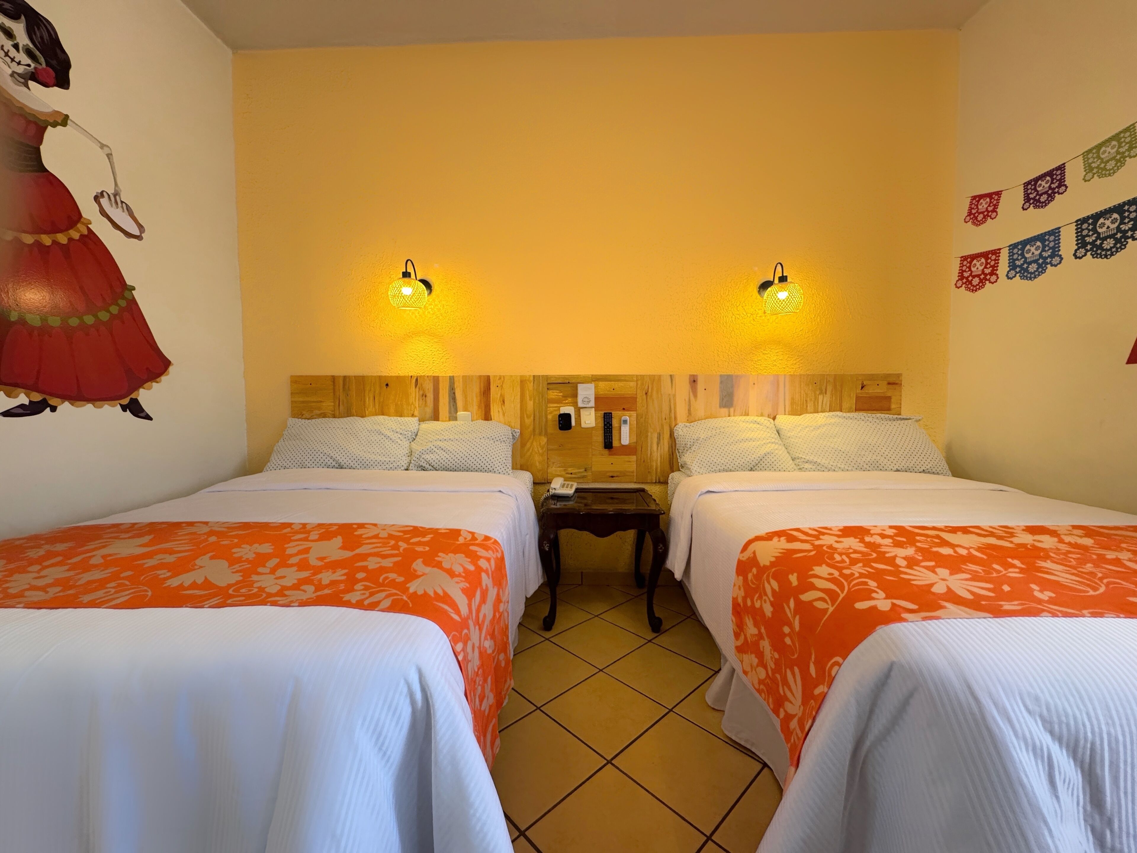 Una camera d'albergo con due letti, ognuno con copriletto arancione e bianco, poggiatesta in legno e lampade ai comodini.