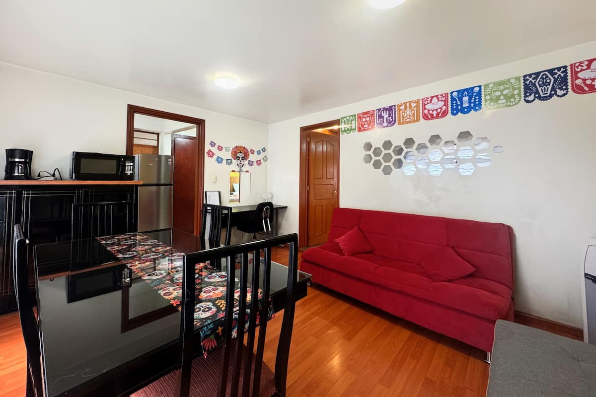 Una sala de estar moderna con un sofá rojo, una mesa de comedor de vidrio y una zona de cocina con microondas y cafetera.