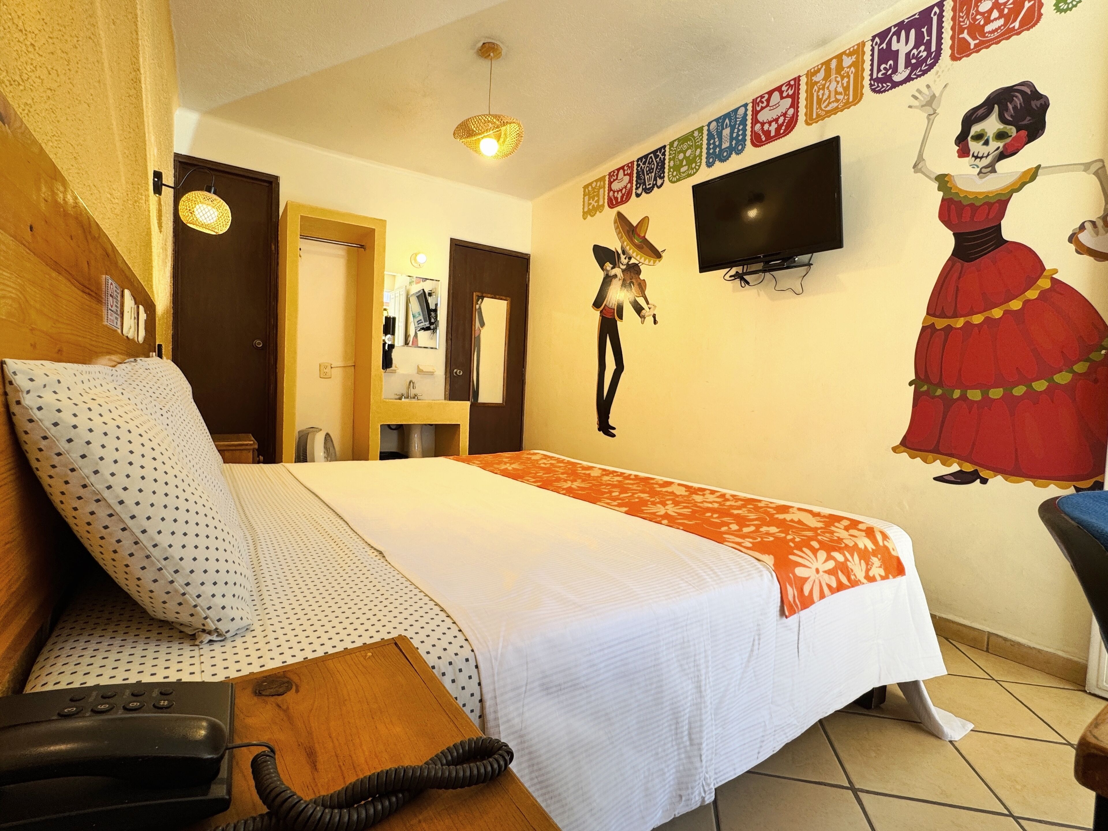 A hotel room with a bed, a telephone, a television, and a wall decorated with cultural artwork.