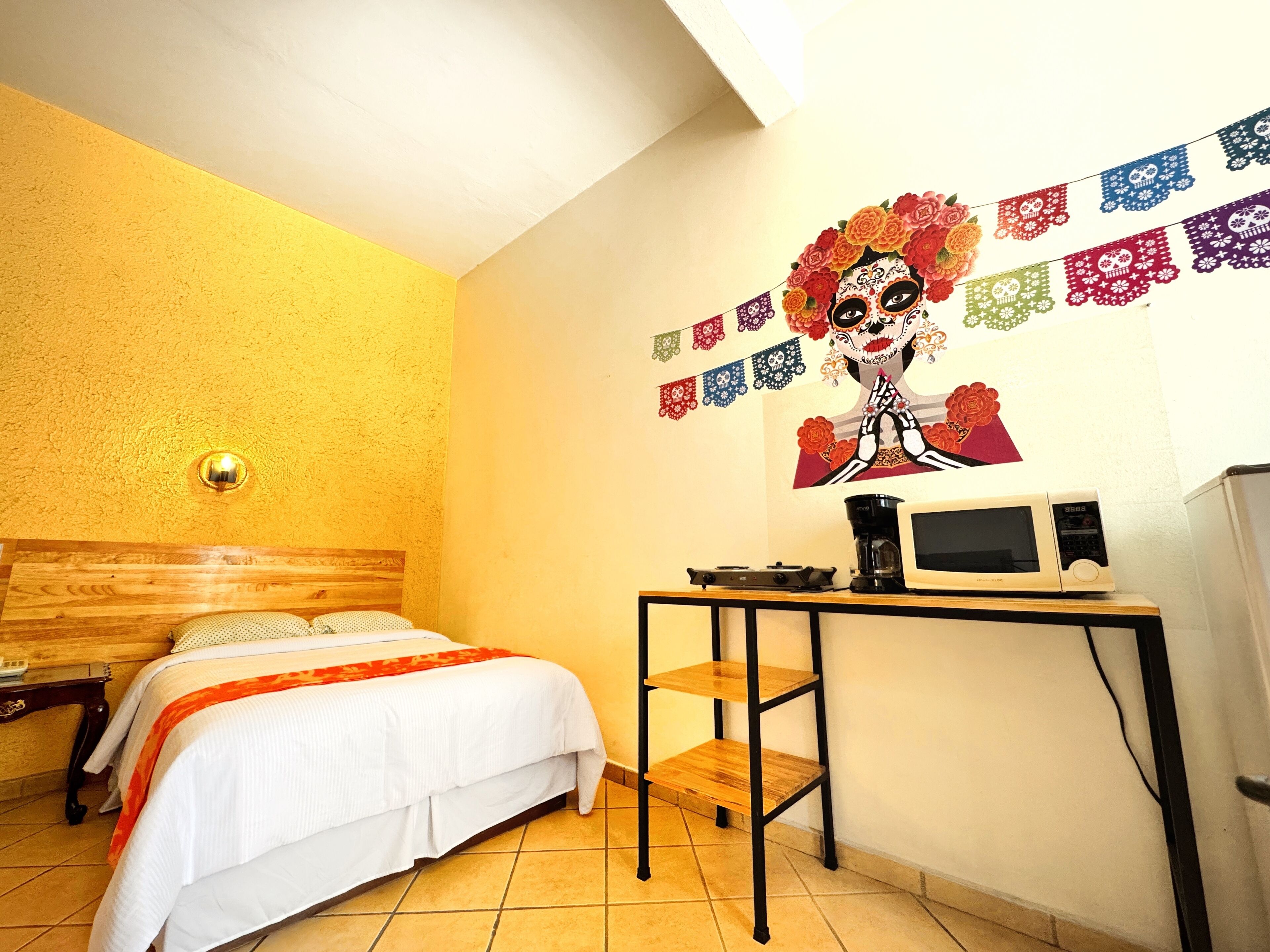Habitación de hotel con cama, escritorio con computadora y cafetera, y una pared decorada con motivos coloridos.