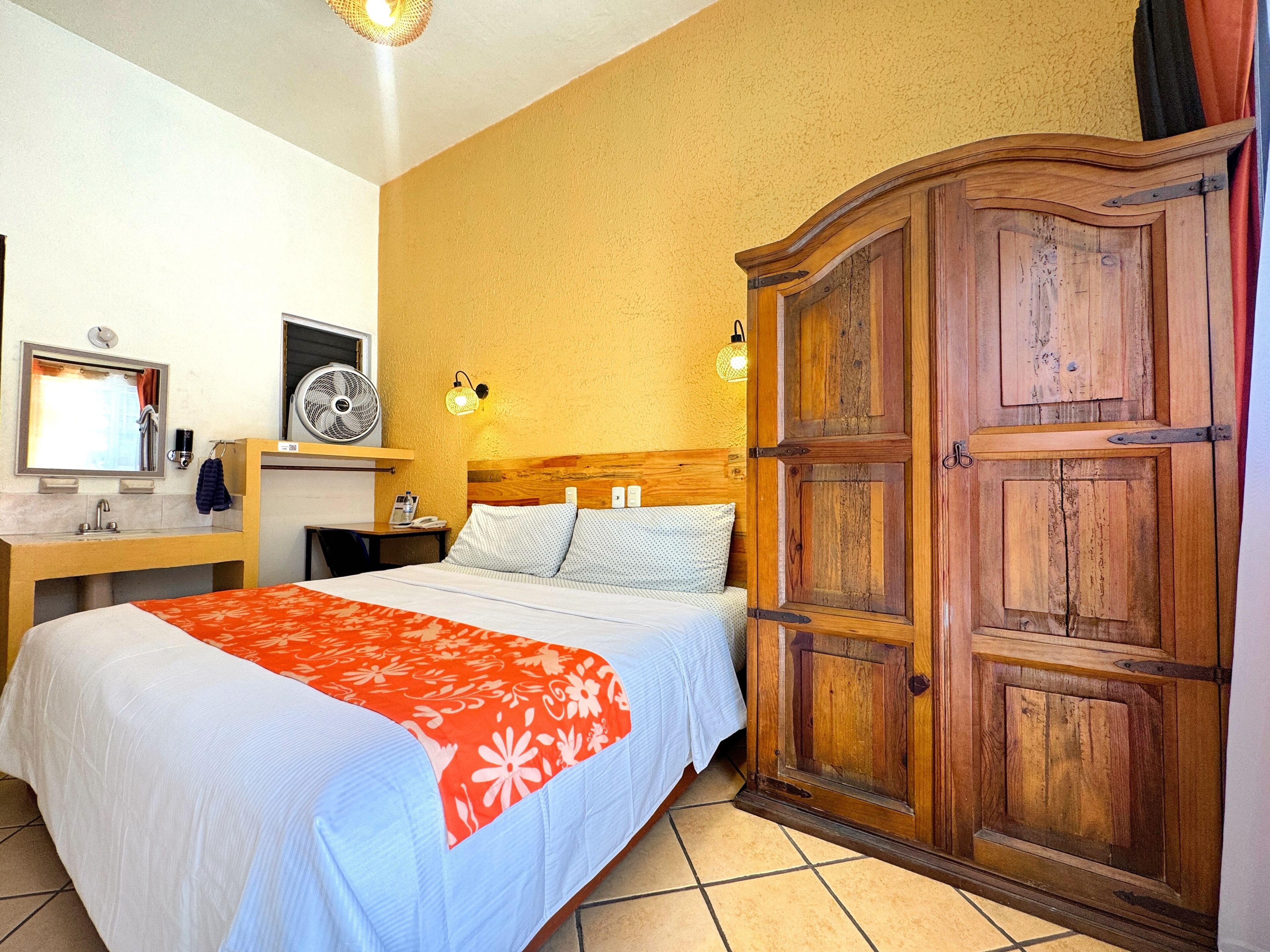 A hotel room with a bed, a wooden wardrobe, a desk with a chair, a mirror, a fan, and a wall-mounted light fixture.