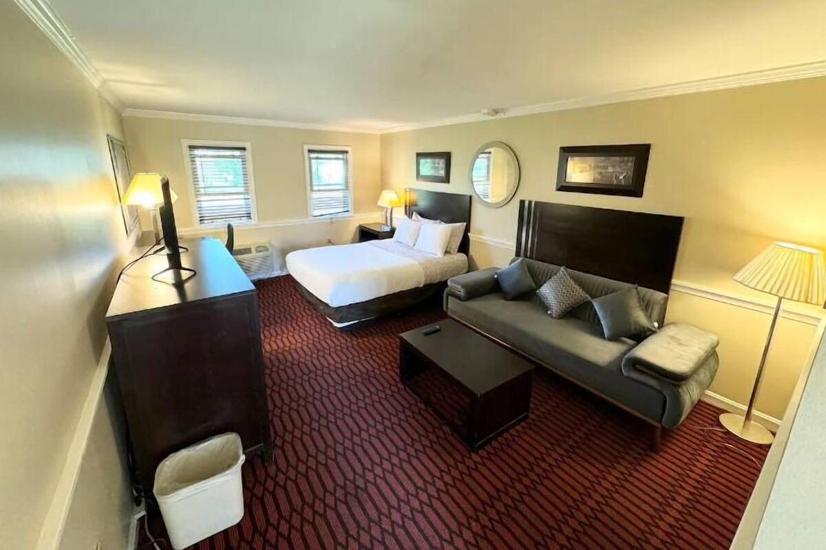 A hotel room with a bed, a sofa, a desk, and a chair. There are two windows with blinds, a lamp, and a mirror on the wall.