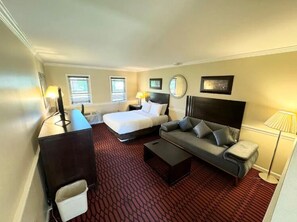 A hotel room with a bed, a sofa, a desk, and a chair. There are two windows with blinds, a lamp, and a mirror on the wall. - Kiteville Lancaster (Strasburg)