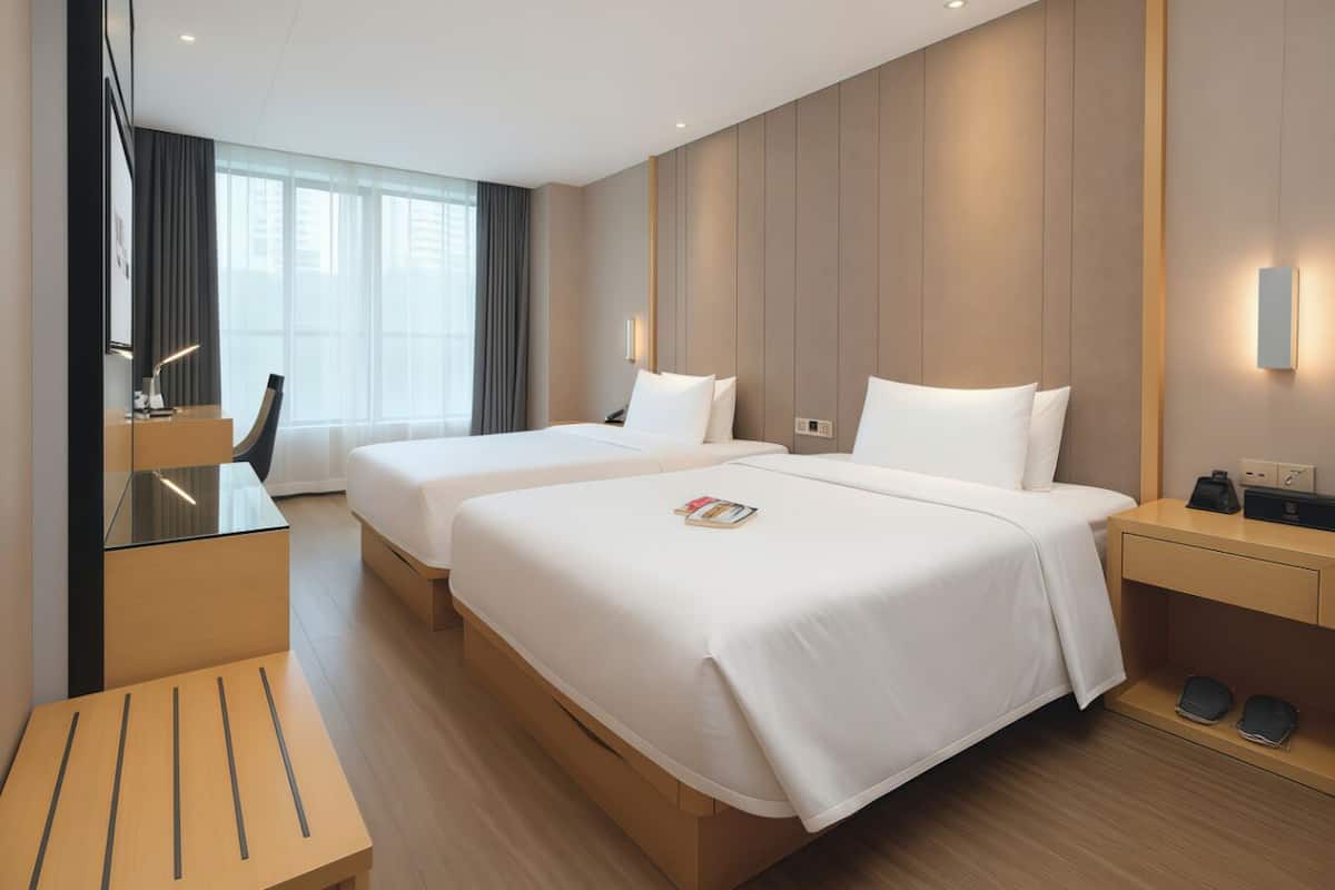 A hotel room with two beds, a desk, a chair, a bedside table, and a large window.