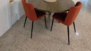 A dining area with a round table, six orange chairs, a ceiling light fixture, and a framed abstract painting on the wall.