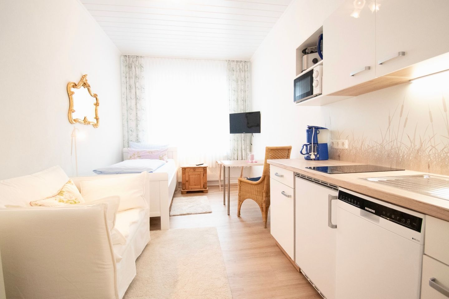 Una habitación moderna y luminosa con un sofá blanco, una pequeña mesa de comedor, una silla de mimbre y una zona de cocina con armarios blancos y un lavavajillas Siemens.