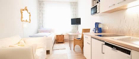 A modern, well-lit room with a white sofa, a small dining table, a wicker chair, and a kitchen area with white cabinets and a Siemens dishwasher.