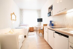 Quarto moderno e bem iluminado com sofá branco, pequena mesa de jantar, cadeira de vime e cozinha equipada com armários brancos e lava-louças Siemens.