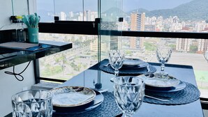 A dining table set with plates, glasses, and cutlery, overlooking a cityscape with buildings and mountains. - Apartamento Novo A 600 Metros DA Praia (Balneario Cidade Atlantica Guarujá)