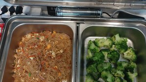 A stainless steel food service station with two compartments: one containing a noodle dish with vegetables and eggs, and the other with steamed broccoli. - Yizhi Hotel Panyu Plaza Shiqiao Subway   (Guangzhou)