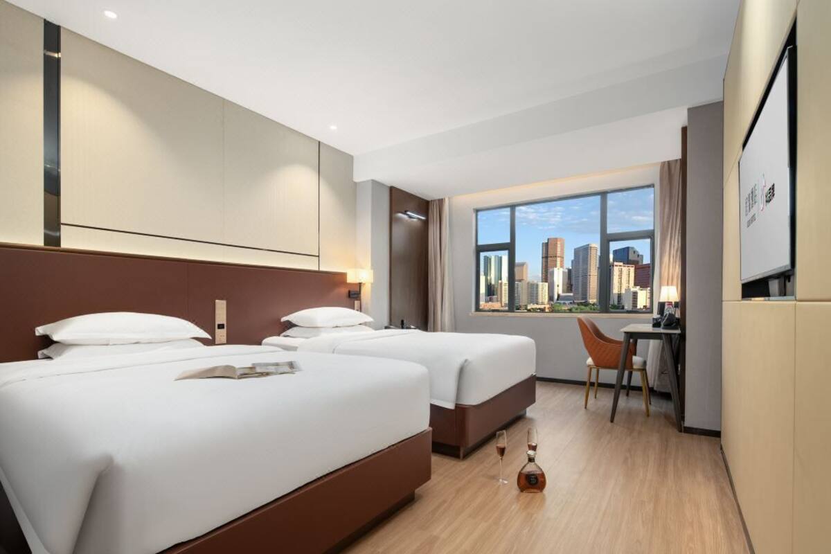 A hotel room with two beds, a desk, and a chair. There is a large window with a city view.