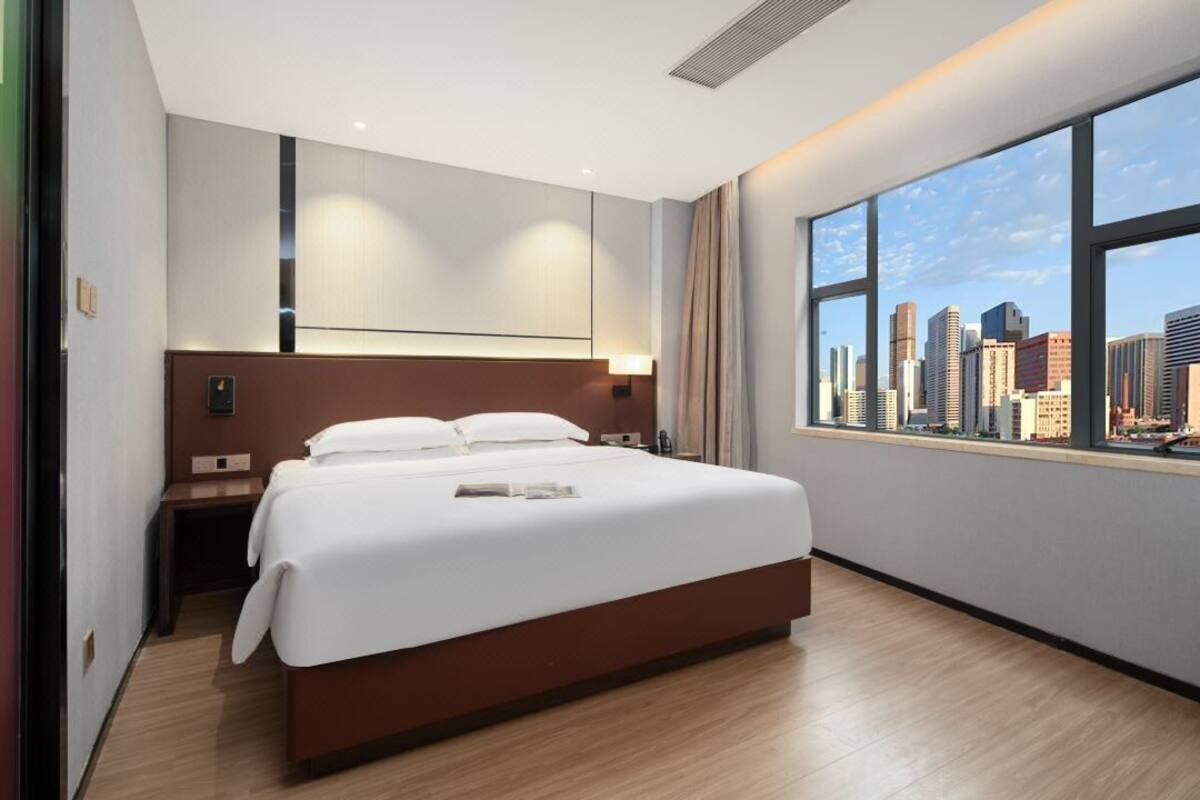 A modern hotel room with a large bed, a nightstand, a desk, and a view of a city skyline through large windows.