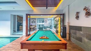 A pool table in a well-lit indoor space with a view of a pool outside. - Villa Rumpi Seminyak by Nagisa Bali (Legian)