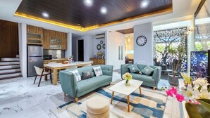 A modern living room with a sofa, ottoman, and coffee table, a dining area with a table and chairs, and a kitchen with wooden cabinets and appliances. - Villa Rumpi Seminyak by Nagisa Bali (Legian)