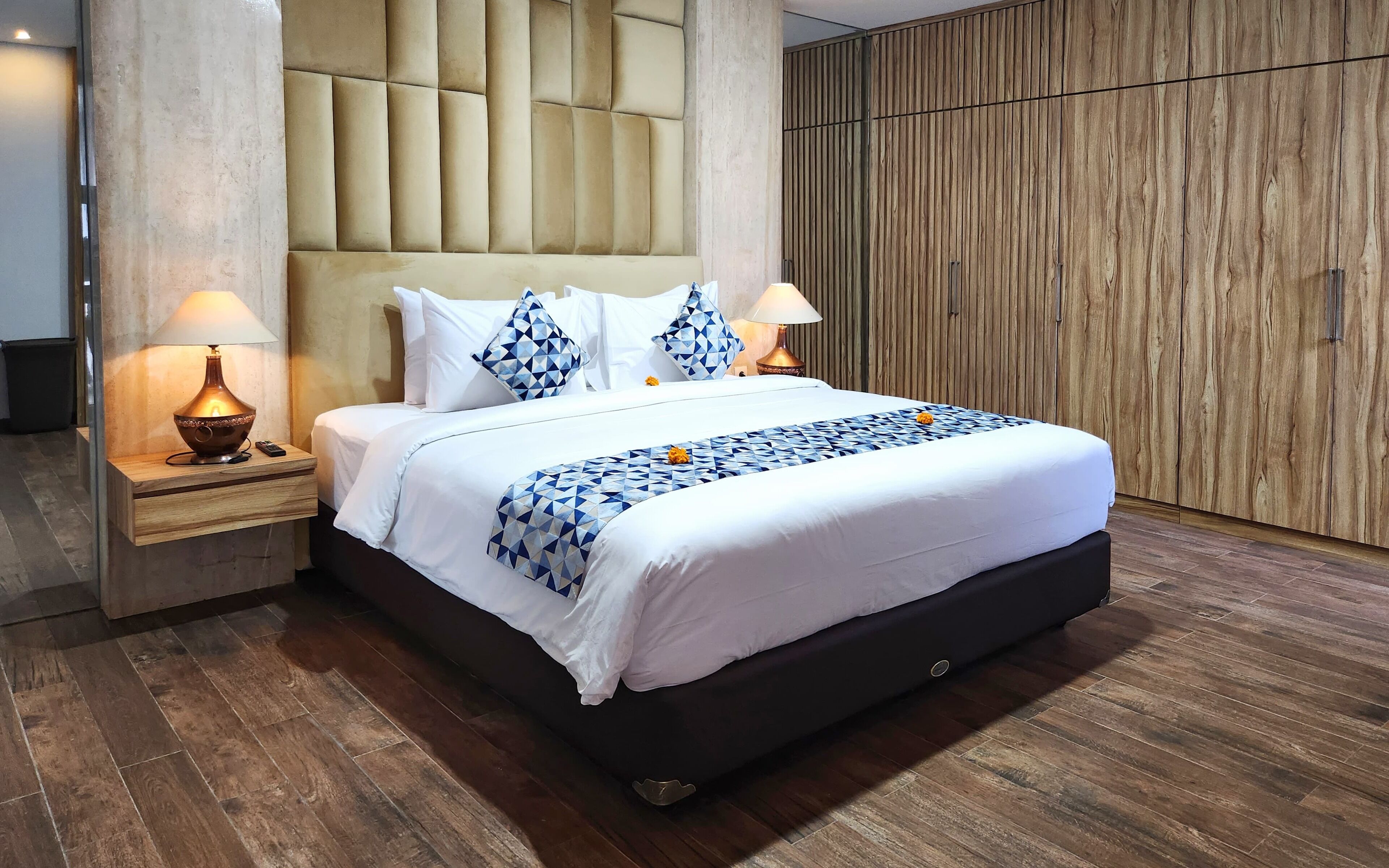 A hotel room with a large bed, two bedside tables with lamps, a wooden wardrobe, and a modern padded headboard.